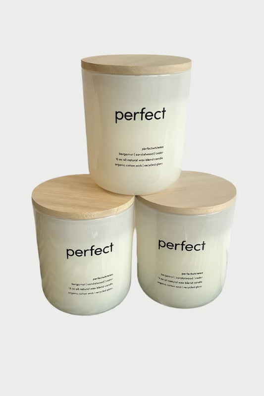 Three "Perfect White Tee" candles by perfectwhitetee in glass jars with wooden lids are arranged in a pyramid formation, with two candles at the base and one on top. Each jar has "Perfect White Tee" printed on it and exudes scents of sandalwood and bergamot, set against a green palm leaf background.