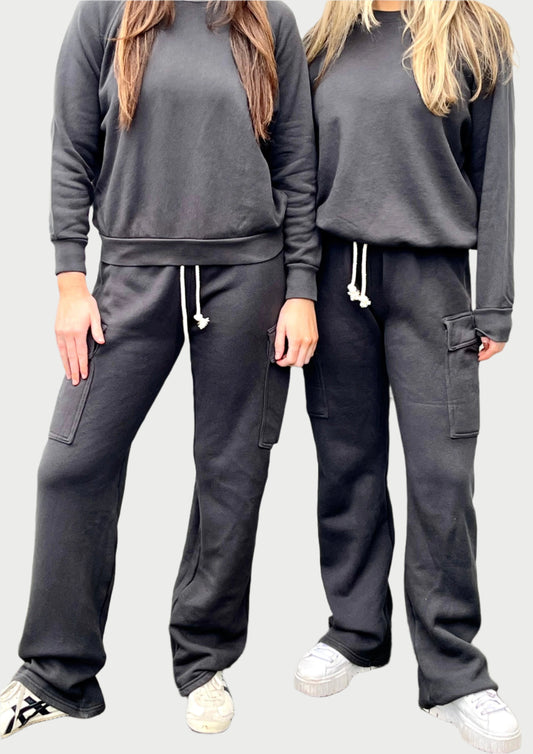 Two people stand side by side wearing matching black sweatsuits with long sleeves and drawstring waist pants. The person on the left has long, dark hair, while the other has long, blonde hair. Both are wearing white sneakers against a light-colored wall, embodying the relaxed style of perfectwhitetee Fleece Cargo Pant.