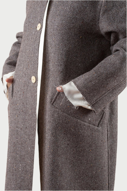 A person exudes an elegant look in the Coat w/scarf by Phisique du role, featuring a brown herringbone pattern, ivory buttons, and white inner cuffs. Their hands rest comfortably in the coat's large side pockets, highlighting its refined design and premium material with a visible rich texture.