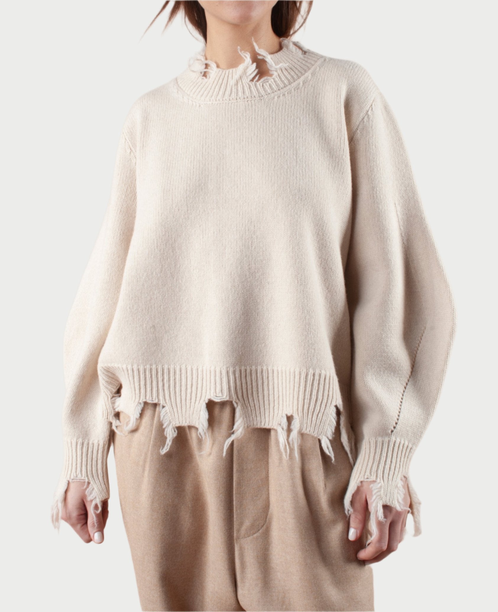 A person is dressed in Phisique du Role's Turnable Sweater with Banana Sleeves, featuring a distressed, frayed hem and cuffs in beige. This stylish sweater is complemented by light brown pants against a plain white background.