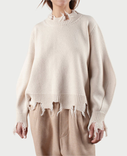 A person is dressed in Phisique du Role's Turnable Sweater with Banana Sleeves, featuring a distressed, frayed hem and cuffs in beige. This stylish sweater is complemented by light brown pants against a plain white background.