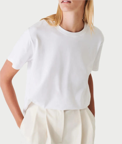 A person wearing a PATRICK ASSARAF Pima Boyfriend Crew T-Shirt in white paired with high-waisted white pants, stands with their hands in their pockets against a softly colored background.