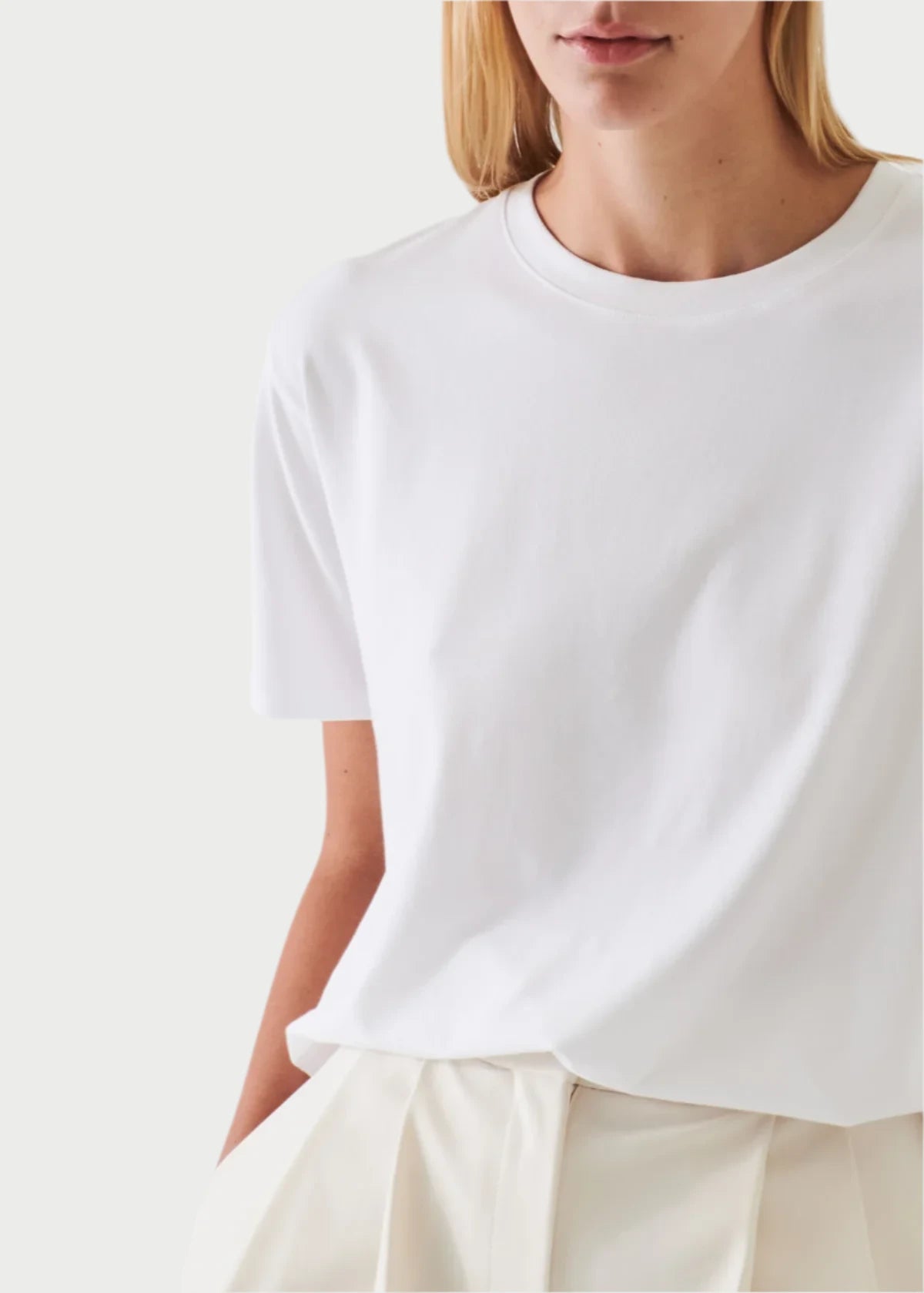 A person wearing the PATRICK ASSARAF Pima Boyfriend Crew T-Shirt in White paired with cream-colored pleated pants stands against a light background. The soft Peruvian Pima cotton fabric ensures comfort, with the focus on the torso, leaving the head partially visible.