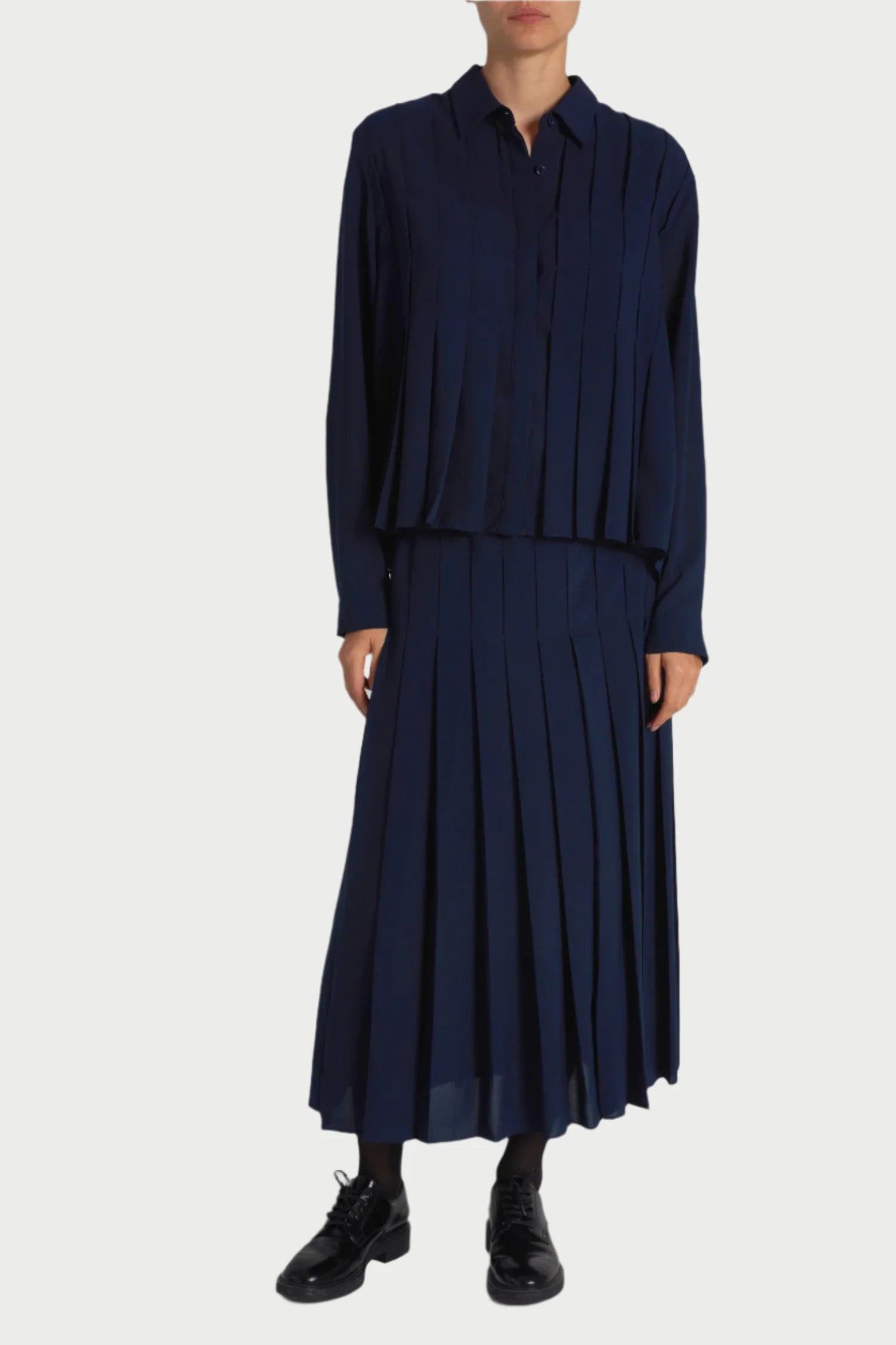 A person is wearing a navy blue, long-sleeve blouse paired with the Saint Art NY Lakelyn Pleated Midi Skirt made from lightweight poly blend fabric. They have black shoes and are standing against a plain white background. Their face is not visible in the image.