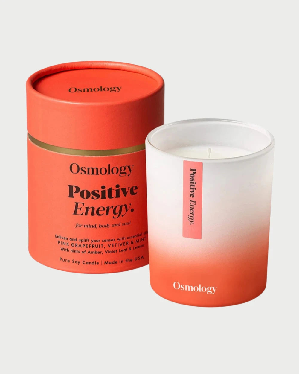 The Osmology Candle, crafted from pure soy, is housed in an orange cylindrical box labeled "Osmology Positive Energy." The white and orange candle features the text "Positive Energy" on its side and emits a scent of pink grapefruit, vetiver, mint, and a touch of patchouli for enhanced aromatherapy benefits.