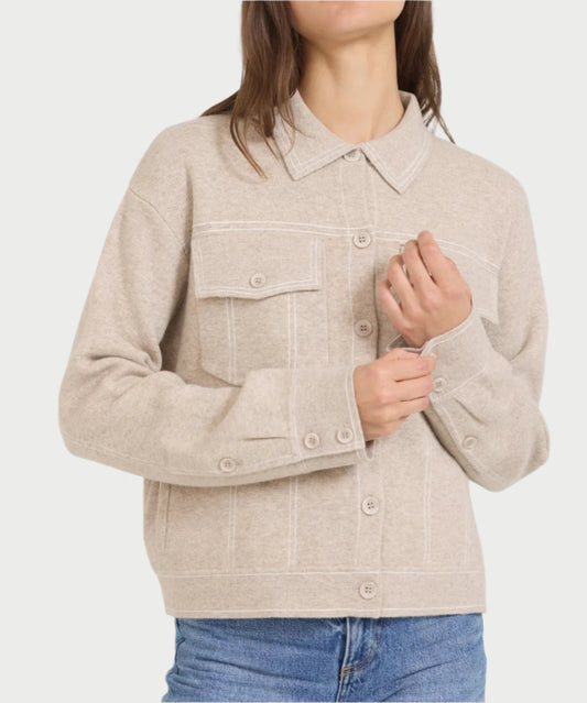 A person wearing the Preston Jacket in Biscuit by One Grey Day, along with blue jeans. The fall jacket, made from a light beige wool and cashmere blend, features two chest pockets, a collar, and buttoned cuffs. The individual is adjusting one of the buttons with their hands.