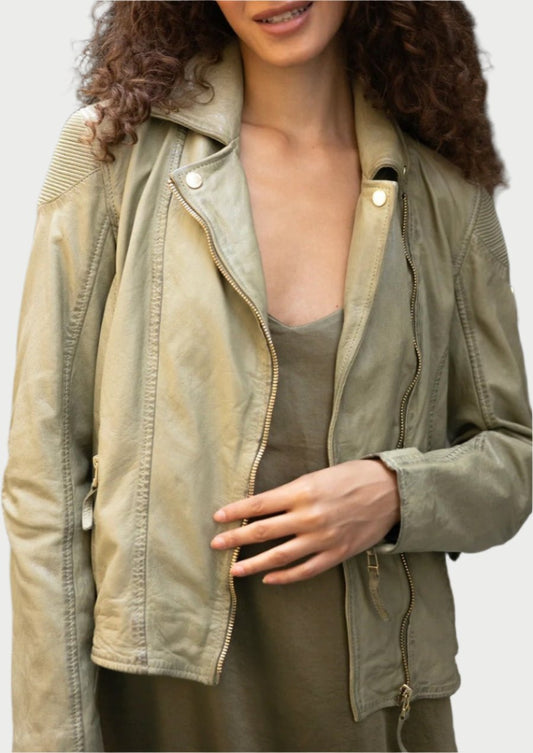 A person with curly hair is wearing a Mauritius Raizel Leather Jacket in sage tone over an olive dress. They are smiling and adjusting the jacket with one hand.