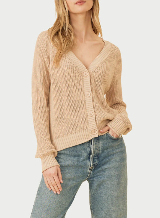 A person with long, wavy blonde hair is wearing the versatile One Grey Day Raleigh Cardigan in beige, featuring a V-neck and buttons down the front. They have paired it with blue jeans and are standing against a plain, light-colored background with a relaxed expression and one hand in their pocket.