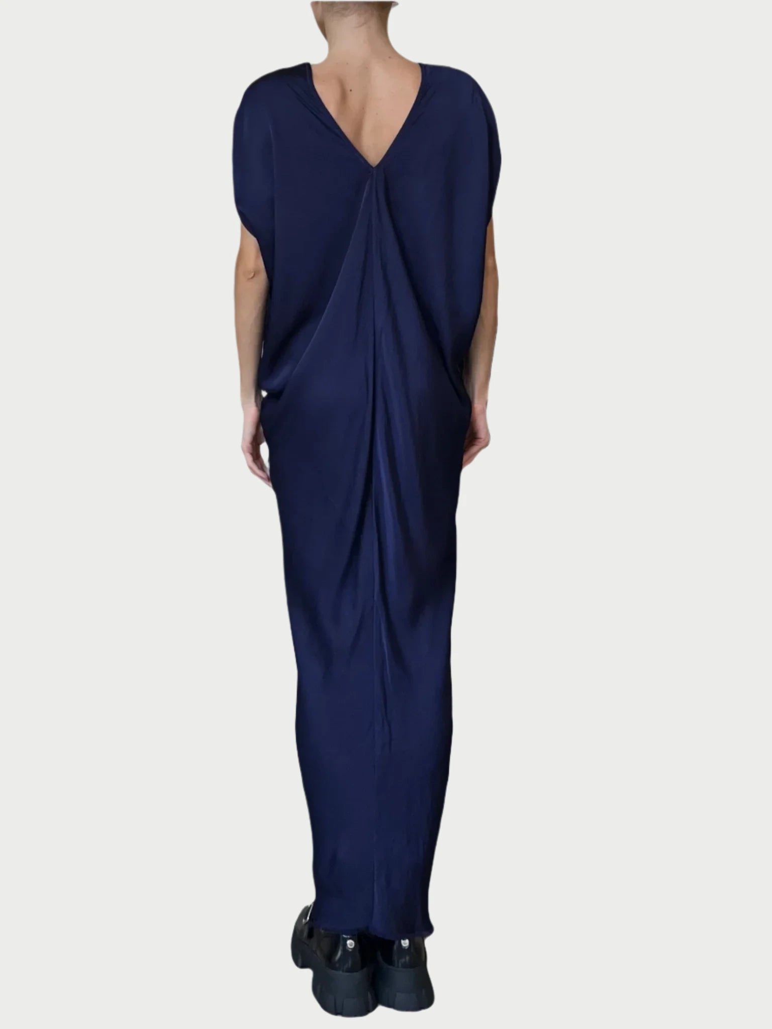 A person is shown from behind, wearing the Leah Dress by Jaga LA. This long, flowing navy blue dress is crafted from high-quality fabric and features a deep V-shaped back. The versatile design drapes elegantly down to the ankles and is paired with black shoes. The plain light gray background adds a subtle sophistication.