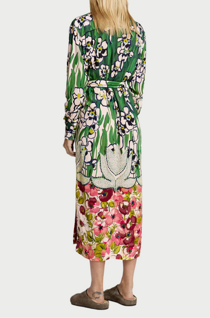 A person in a Smythe Tailored Shirt Dress featuring an Eco Vero-certified, colorful garden print with long sleeves and a belt stands back to the camera. The dress boasts green, pink, and white floral designs. Paired with slippers, the ensemble radiates effortless elegance.