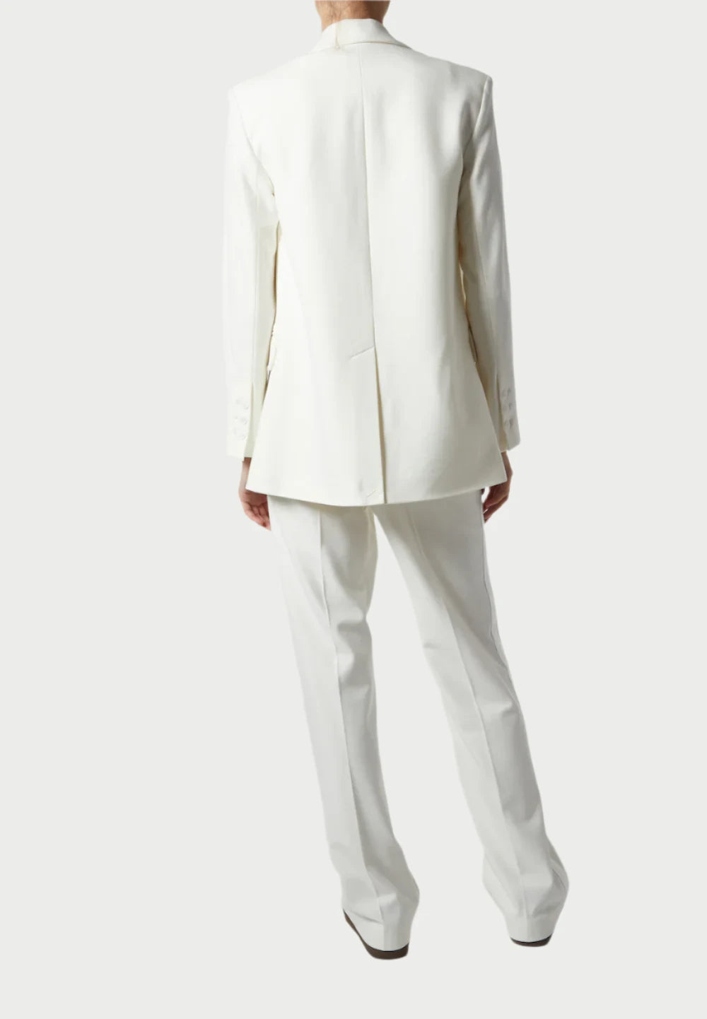 A person showcases the elegance of formalwear in an ivory Dylan Tuxedo Jacket by Saint Art NY, featuring a charmeuse satin lapel, standing against a plain background. The tailored design includes a long jacket and straight pants, exuding sophistication.