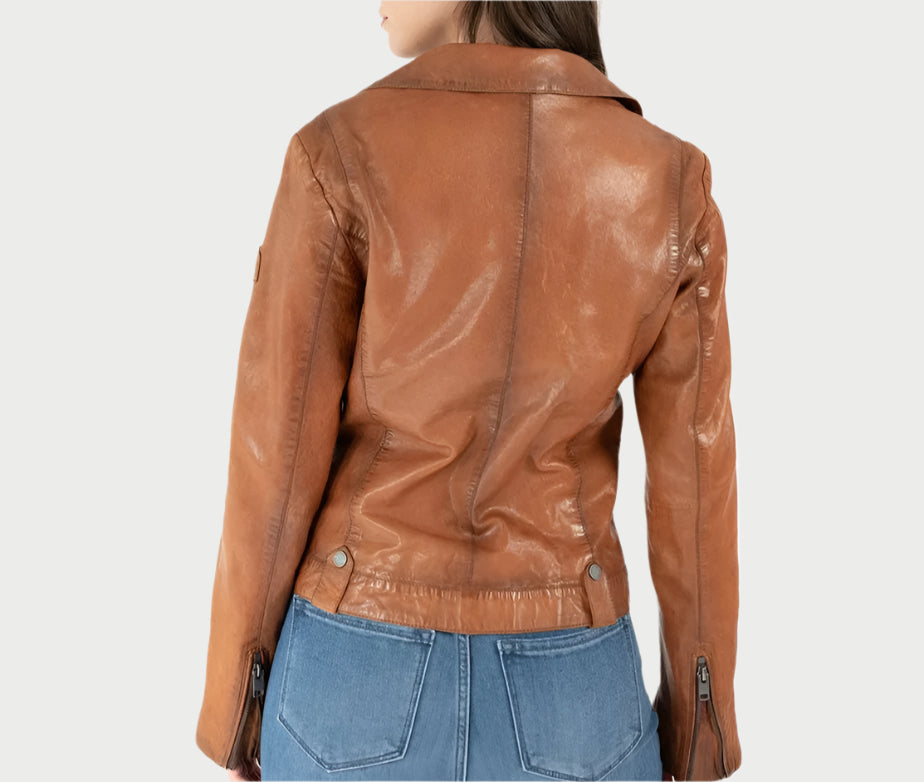 A woman in a cognac-colored Mauritius Fayola Moto leather jacket and blue jeans is seen from behind against a plain background. The jacket features a back seam and sleeve zippers, making it an ideal addition to your Spring closet.