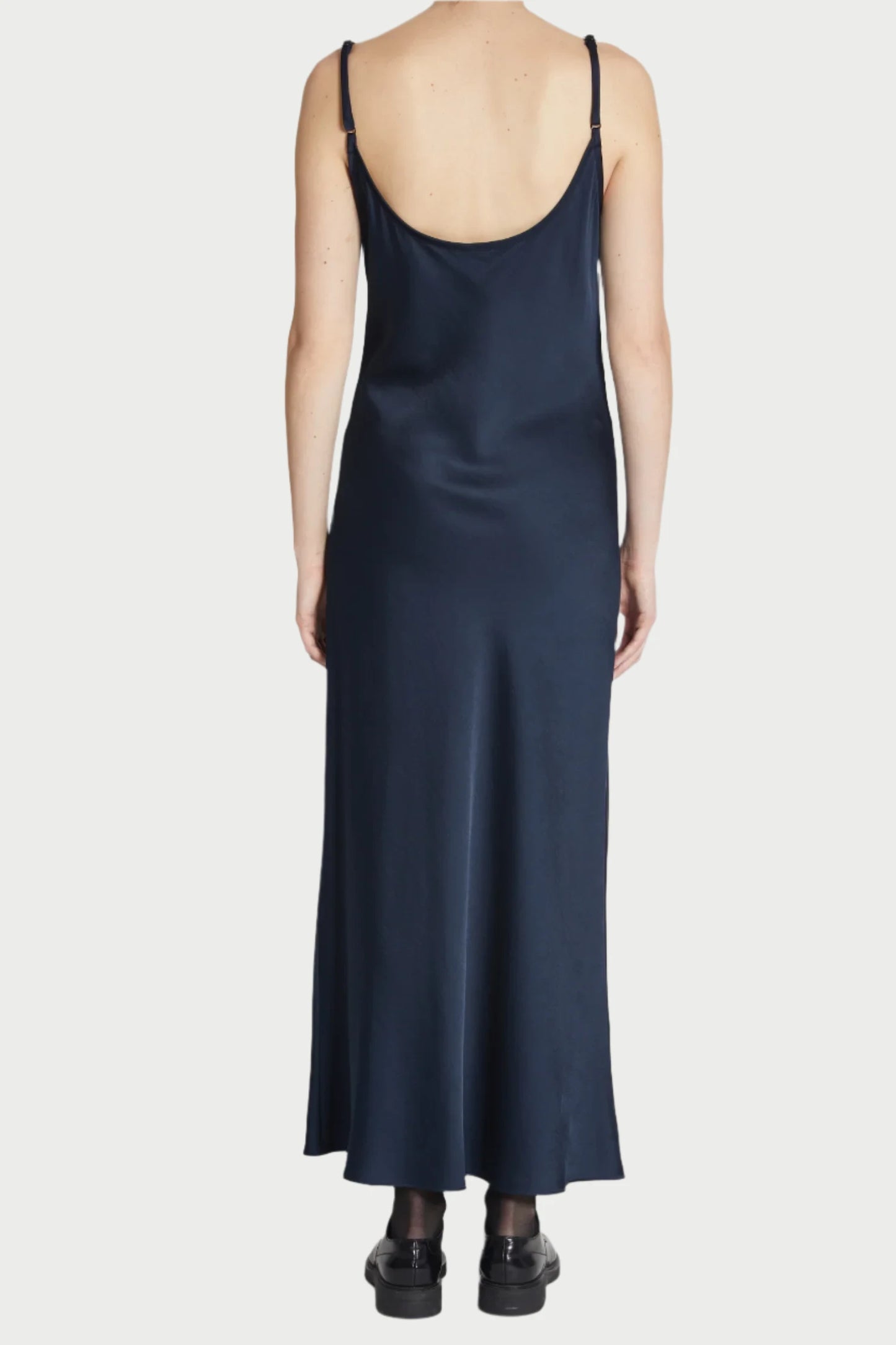 A person is standing with their back facing the camera, wearing the Navy Haley Slip Dress by Saint Art NY. The full-length dress, crafted from heavyweight charmeuse satin in dark navy blue, falls elegantly to their ankles. They are also wearing black ankle socks and black slip-on shoes against a plain light-colored background.