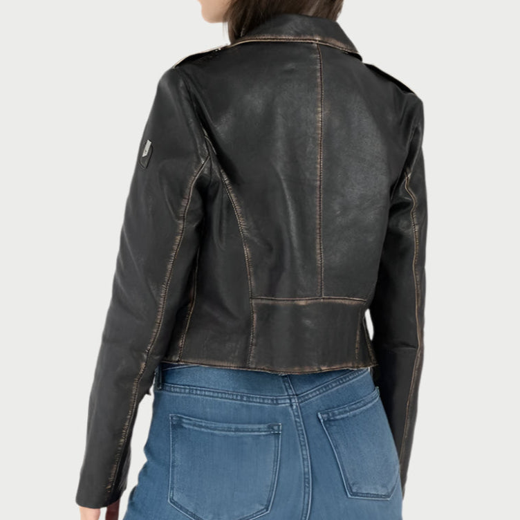 A person wearing the Sharon Moto Jacket by Mauritius, featuring vintage-look black leather with visible stitching and a sleeve patch, paired with blue jeans. They're facing away to highlight the back of the jacket and jeans against a plain light gray backdrop.