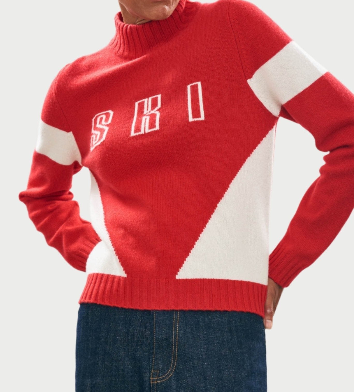 A person wearing the JUMPER Contrast Ski Sweater, showcasing bold red and white color blocks with "SKI" across the chest. The cashmere piece features a stylish turtleneck. Paired with blue jeans, they pose confidently with hands on hips against a white background.