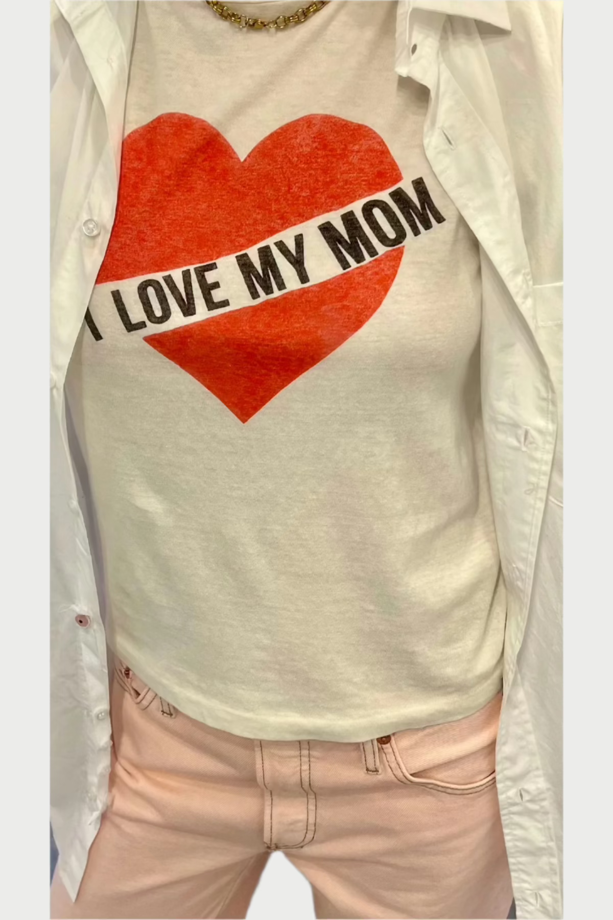 The individual is wearing a snug I Love My Mom T-Shirt by RE/DONE, crafted from 100% cotton and adorned with a red heart and the phrase "I LOVE MY MOM." It’s stylishly layered under an open white jacket and matched with fashionable light pink pants.