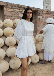 A woman wearing a loose fit white summer Ella Dress by Rhode stands in front of a pile of large pottery jars and a brick wall. She has long black hair and looks directly at the camera, her dress cinched with a braided belt. Behind her, a man in traditional white clothing is arranging the jars.