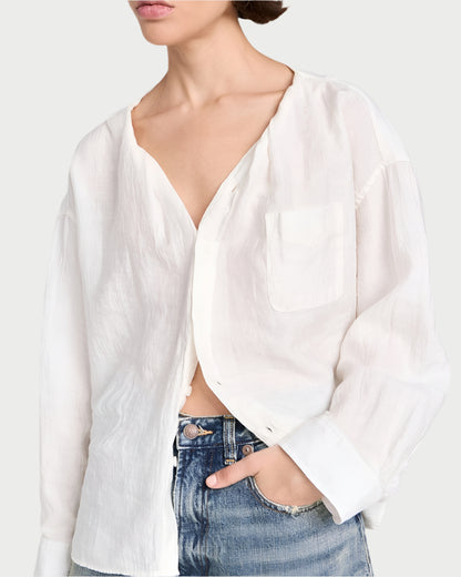 The individual wears R13's Twisted Neck Shirt, featuring a sheer white design with an open collar and long sleeves, crafted from Japanese crinkle lyocell. It includes a chest pocket and effortlessly pairs with faded blue jeans as their hand rests playfully in the jeans' pocket against a neutral background.