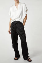 Load image into Gallery viewer, A person wearing a white, short-sleeved V-neck top and high-waisted, cargo-inspired black Saint Art NY Charmeuse Pants with belt and zipper details is standing against a plain background. With one hand in their pocket and sporting black sandals, the outfit exudes a sleek Saint Art NY vibe.