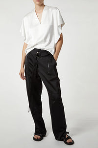 A person wearing a white, short-sleeved V-neck top and high-waisted, cargo-inspired black Saint Art NY Charmeuse Pants with belt and zipper details is standing against a plain background. With one hand in their pocket and sporting black sandals, the outfit exudes a sleek Saint Art NY vibe.