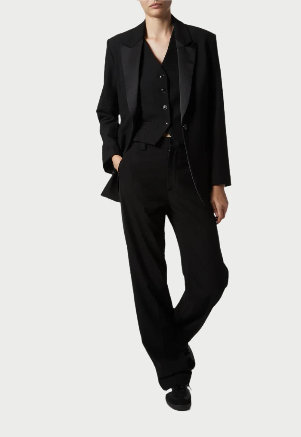 The individual stands poised in a debonair black three-piece suit, featuring a Saint Art NY Dylan Tuxedo Jacket with charmeuse satin lapels. Their hands rest by their sides, complemented by polished black shoes against a light background.