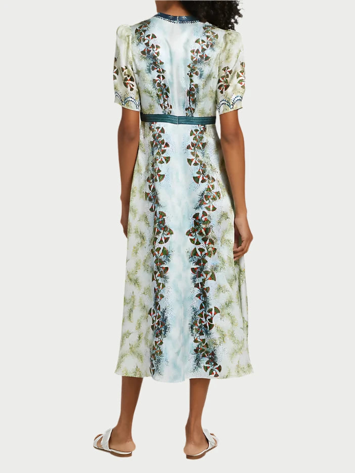 A person with curly hair is shown from the back, wearing the Saloni Tabitha Midi Dress with a blue and green botanical print. Perfect for a warm-weather celebration, the dress features short puff sleeves, a cinched waist with a black band, and falls below the knees. They are also wearing white slip-on sandals.