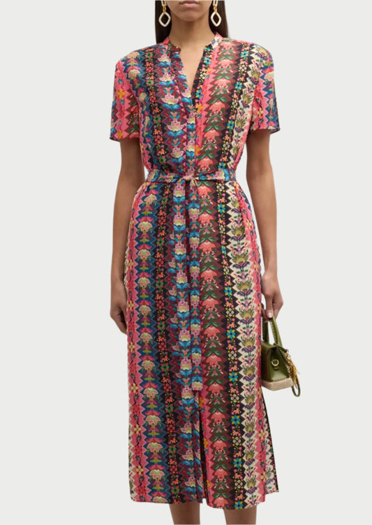 A person is wearing a vibrant, multi-colored floral Vicki Dress with short sleeves and a belted waist, showcasing contemporary silhouettes by Saloni. They hold a small green handbag in their left hand and have long hair. The background is plain white.