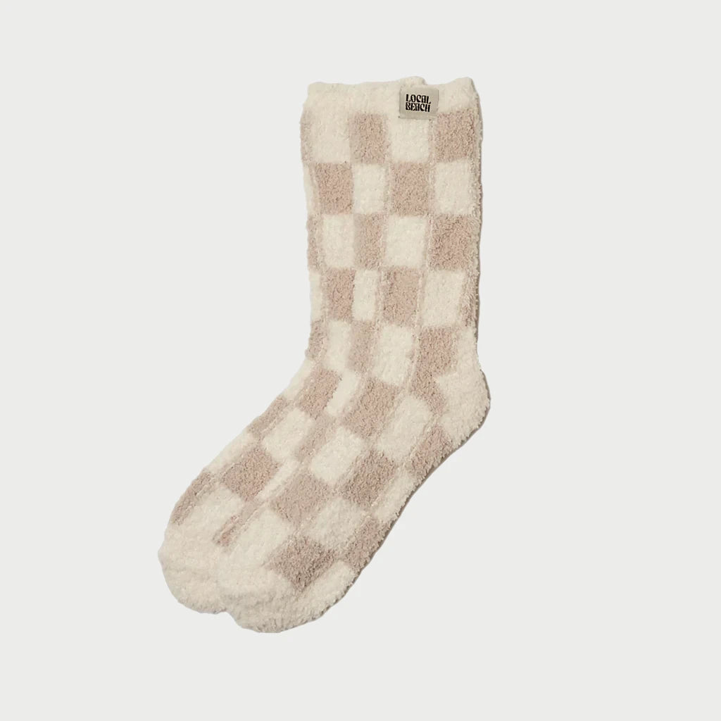 Introducing a stylish piece from LOCAL BEACH’s Cozy Socks lineup, this individual slipper sock features a chic beige and white checkerboard design accompanied by a small tag near the top. Made with Feather Knit construction, it is presented against a simple white background.