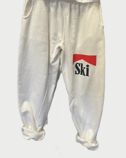 Introducing the "Sweatpants w/Ski print" by SUPER READY: These white sweatpants feature the word "Ski" prominently displayed in black and red on one leg. Designed with an elastic waistband and rolled-up cuffs, they are perfect for hitting the slopes or lounging in style.