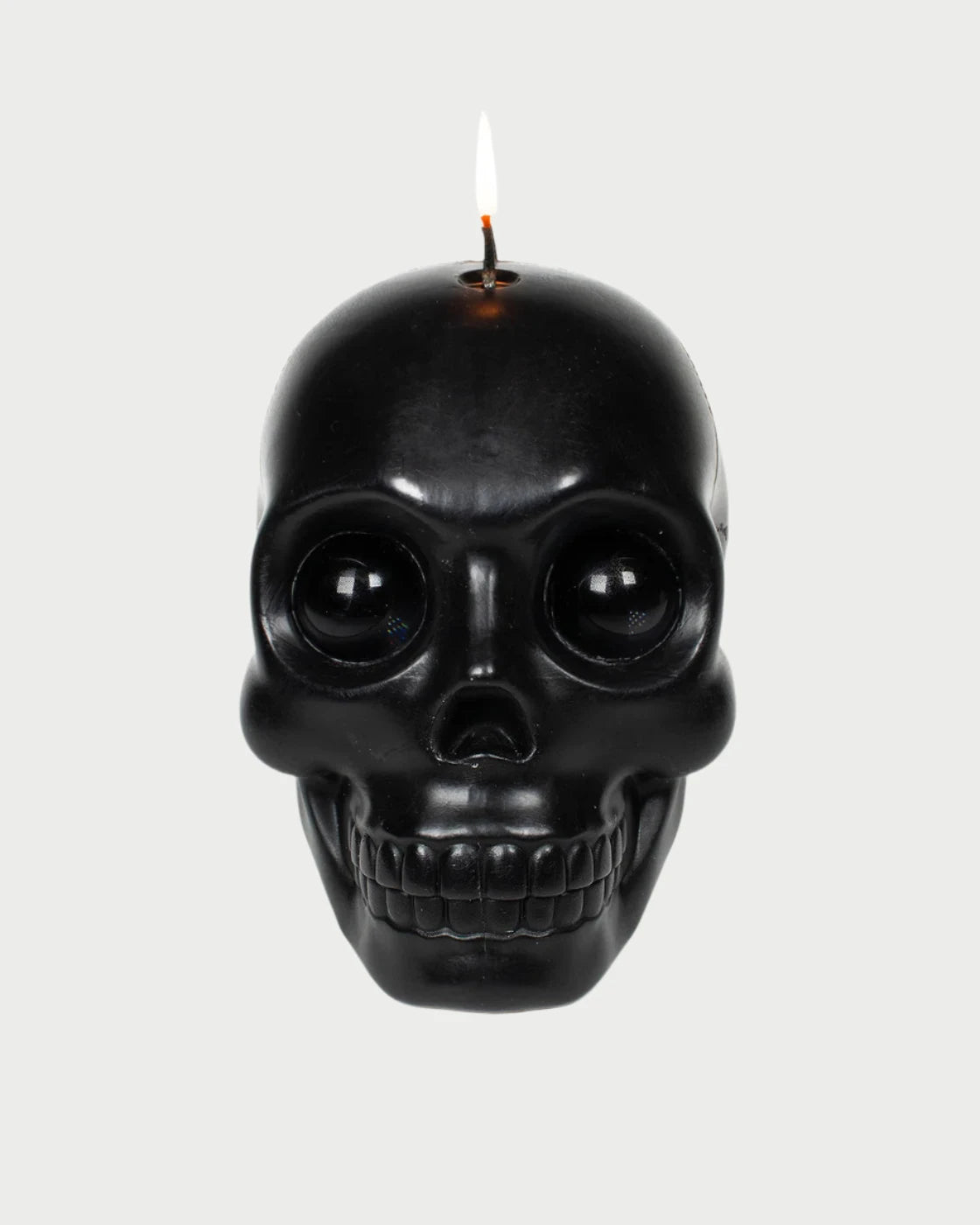 The Skull Candle by 54° Celsius, crafted from paraffin wax and shaped like a black skull, is illuminated at the top, casting a glow against a plain light background. It features intricate facial contours, crystal eyeballs in its hollow eyes, and a toothy grin.