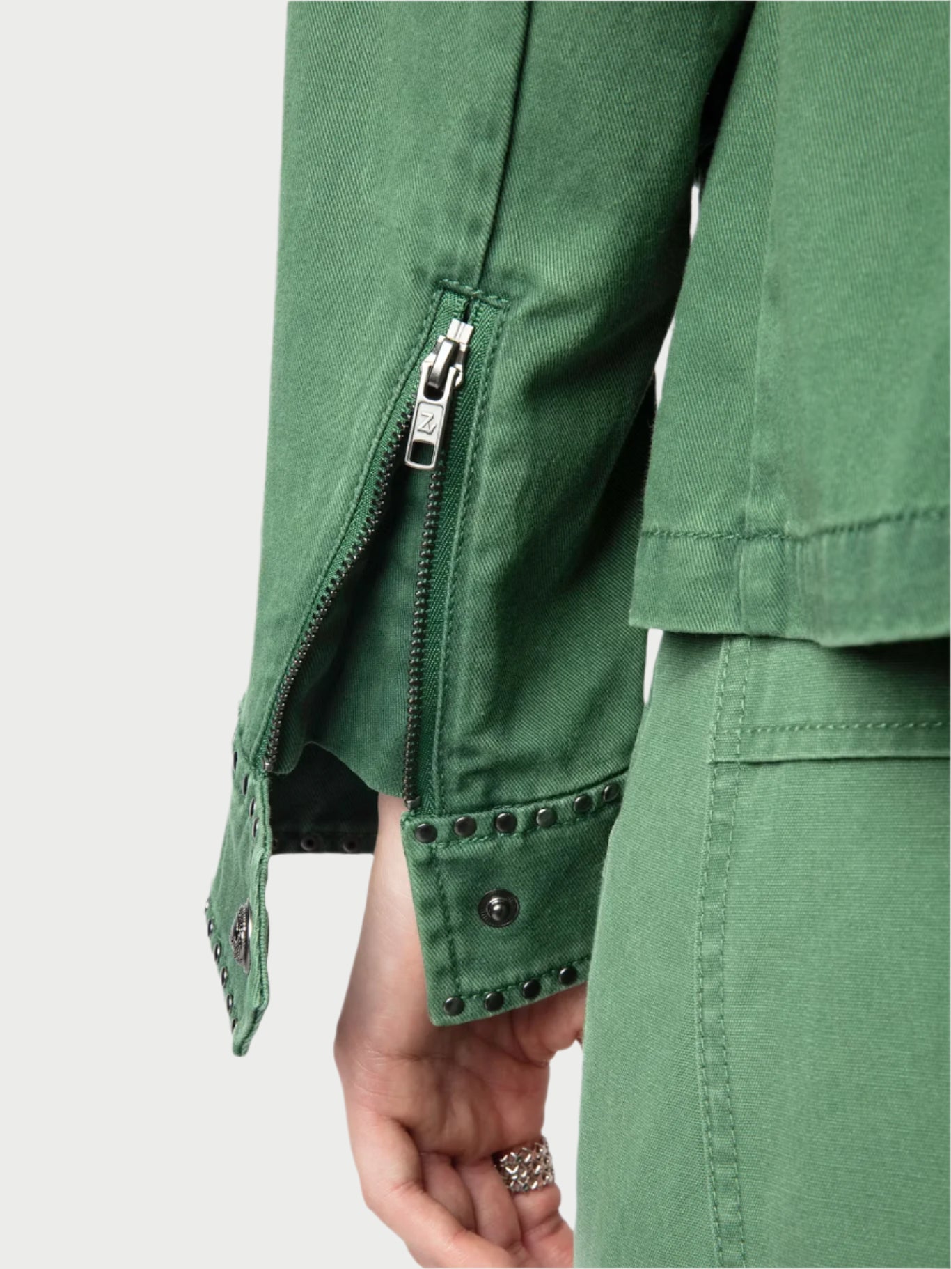 Close-up of someone wearing an army green Klimi Utility Jacket by Zadig & Voltaire with visible zipper and studded cuff details, alongside cotton denim pants. Their hand, decorated with rings, is partially visible.