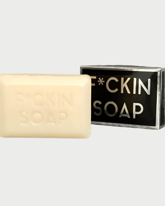 In front of its packaging, a deeply moisturizing beige bar of F*cking Bar Soap by Rude Dude is showcased. The black packaging prominently features bold white text that reads "F*CKIN SOAP," which is also embossed on the bar itself, enhanced with a subtle champagne fragrance.