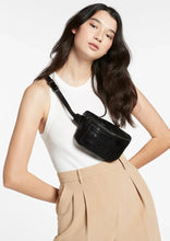 Load image into Gallery viewer, A woman with long, dark hair wears a white sleeveless top and beige high-waisted pants. She has a black Status Anxiety Belt Bag made of lamb skin leather with gunmetal hardware slung over her shoulder. She is standing against a plain background.