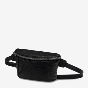 A black lamb skin leather Belt Bag with a textured surface by Status Anxiety. The bag features gunmetal hardware, a main compartment with a zipper closure, an adjustable strap, and a smaller front pocket also with a zipper closure. The background is plain white.