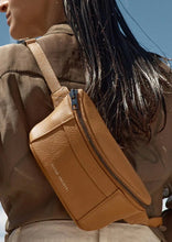 Load image into Gallery viewer, A person with long dark hair is shown from the back, wearing a light brown Status Anxiety Belt Bag with gunmetal hardware secured around their shoulder. The person&#39;s outfit appears to be a sheer, brown top. The background is a clear blue sky.