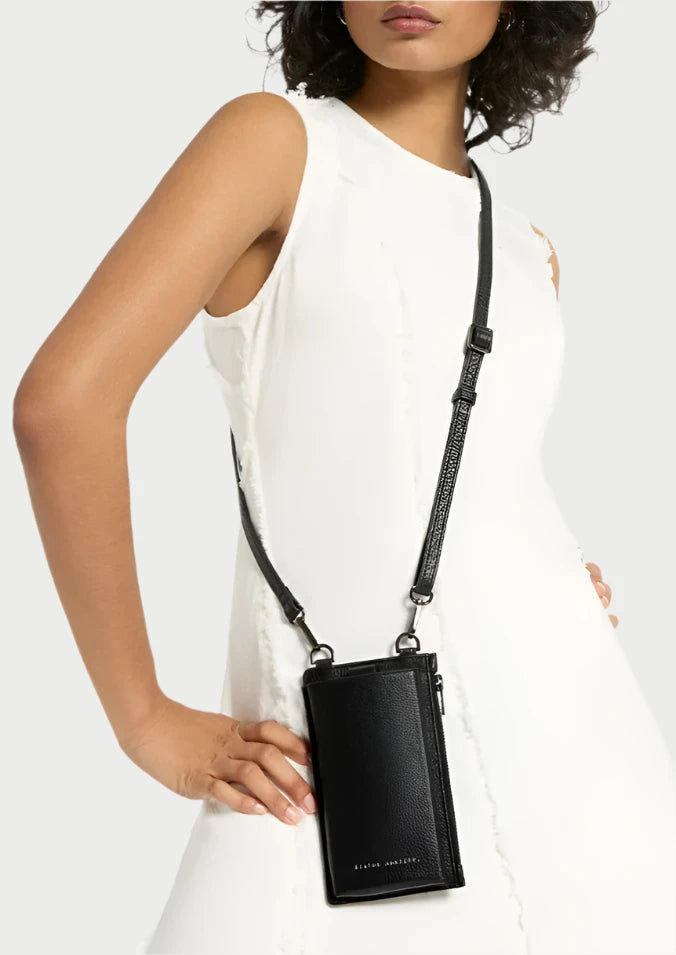 A woman in a sleeveless white dress stands with one hand on her hip, showcasing a black Voyager Phone Bag by Status Anxiety with an adjustable strap. The bag has a sleek design and is worn across her body like a stylish phone case. The background is plain white.