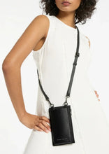 Load image into Gallery viewer, A woman in a sleeveless white dress stands with one hand on her hip, showcasing a black Voyager Phone Bag by Status Anxiety with an adjustable strap. The bag has a sleek design and is worn across her body like a stylish phone case. The background is plain white.