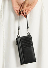 Load image into Gallery viewer, A voyager is holding a black Status Anxiety Voyager Phone Bag with a wrist strap. The bag, reminiscent of a sleek phone case, features multiple card slots on the front and a zippered compartment. The background is a light fabric, possibly part of the person&#39;s clothing.