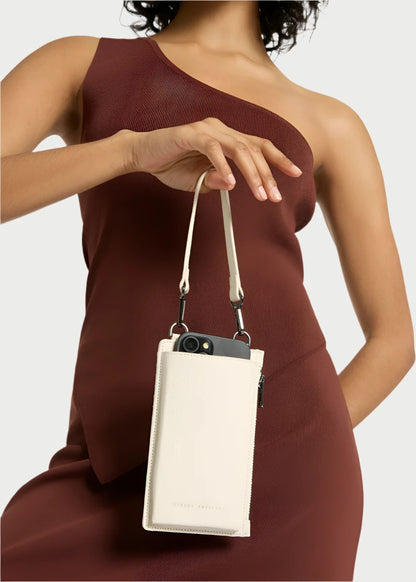 A person in a one-shoulder, sleeveless brown dress holds a cream-colored Voyager Phone Bag by Status Anxiety with a smartphone partially visible inside. The background is plain white.