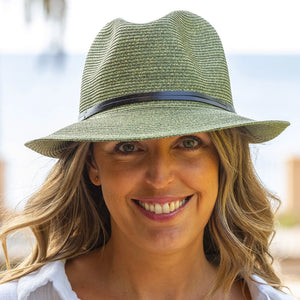A woman with long, wavy blonde hair is wearing a green fedora hat with a black band and a white top. She is smiling warmly, showcasing one of Travaux en Cours' Summer Hats. The background is slightly blurred, suggesting an outdoor setting, possibly near a beach or waterfront.