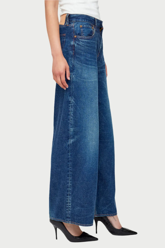 A person dressed in the high-waisted Moussy Emporia Super Wide Leg Jeans, crafted from 100% cotton, paired with a white top and black pointed high-heel shoes, standing in a side profile against a plain light background.