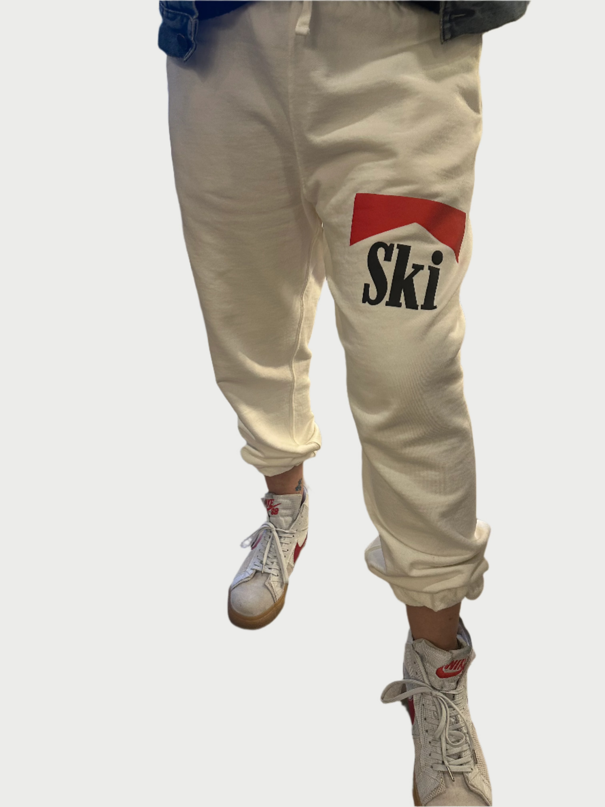 A person dressed in SUPER READY's Sweatpants w/Ski print, showcasing a red and black "Ski" logo on the side, stands poised in their stylish white high-top sneakers with red accents against a plain backdrop.