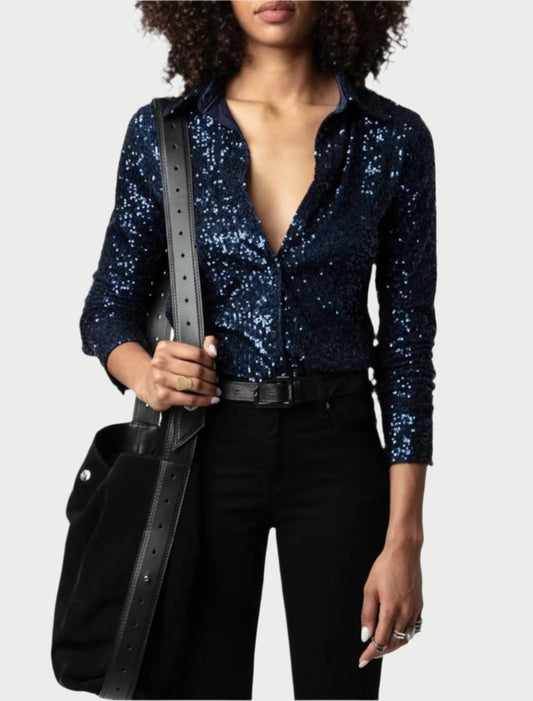 A person dressed in a Tawny Sequin Top from Zadig & Voltaire, paired with black pants and a chic black belt, holds a large black bag featuring studded details. Adorned with rings on their fingers, they exude elegance against the plain background.