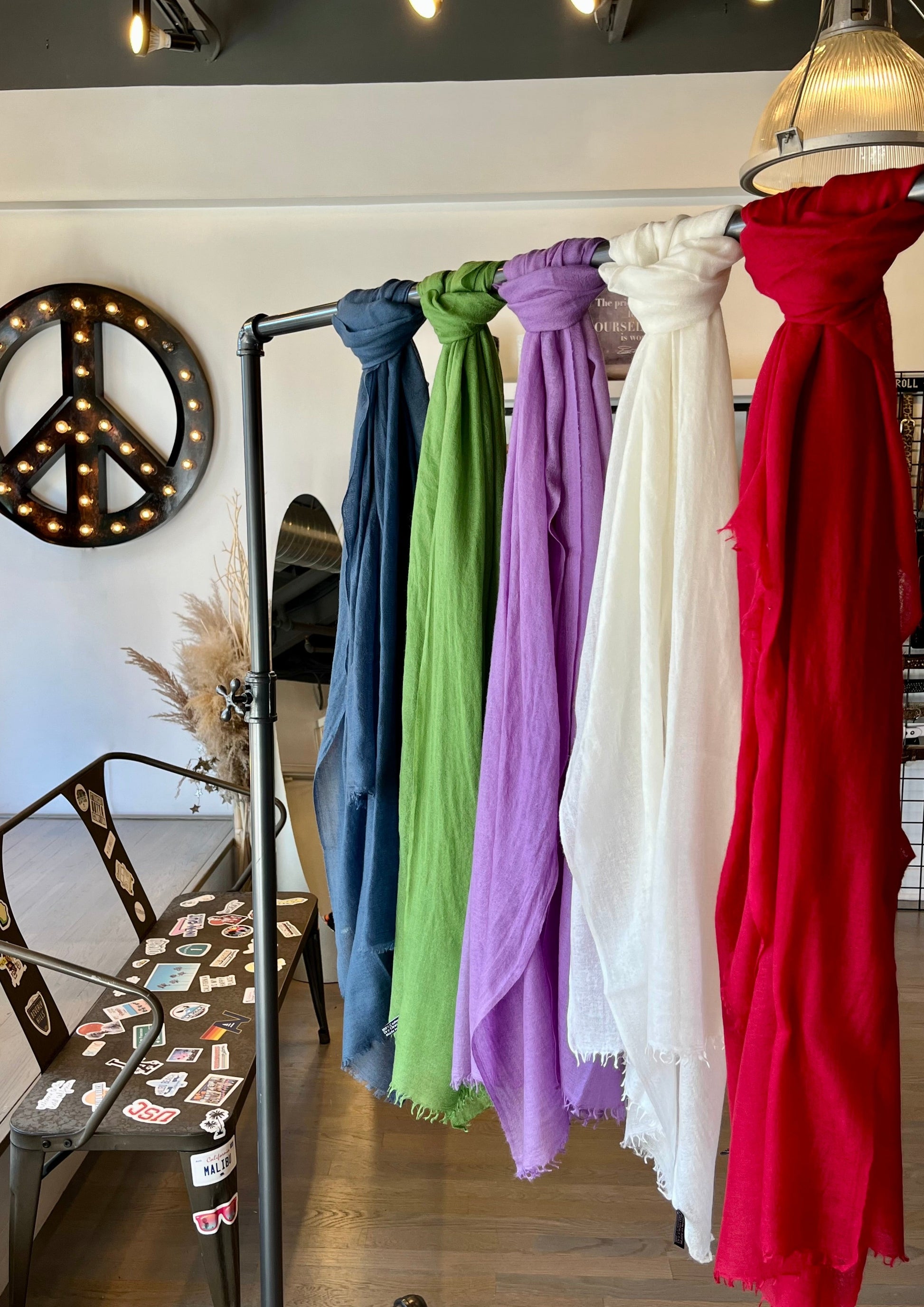 A display of colorful Whisperweight Cashmere Scarves by Meg Cohen hanging on a metal rack inside a stylish café. The scarves in blue, green, purple, white, and red are displayed in a row. There is a peace sign wall decoration and a table with chairs in the background. Luxurious hats made from natural fibers are also available.