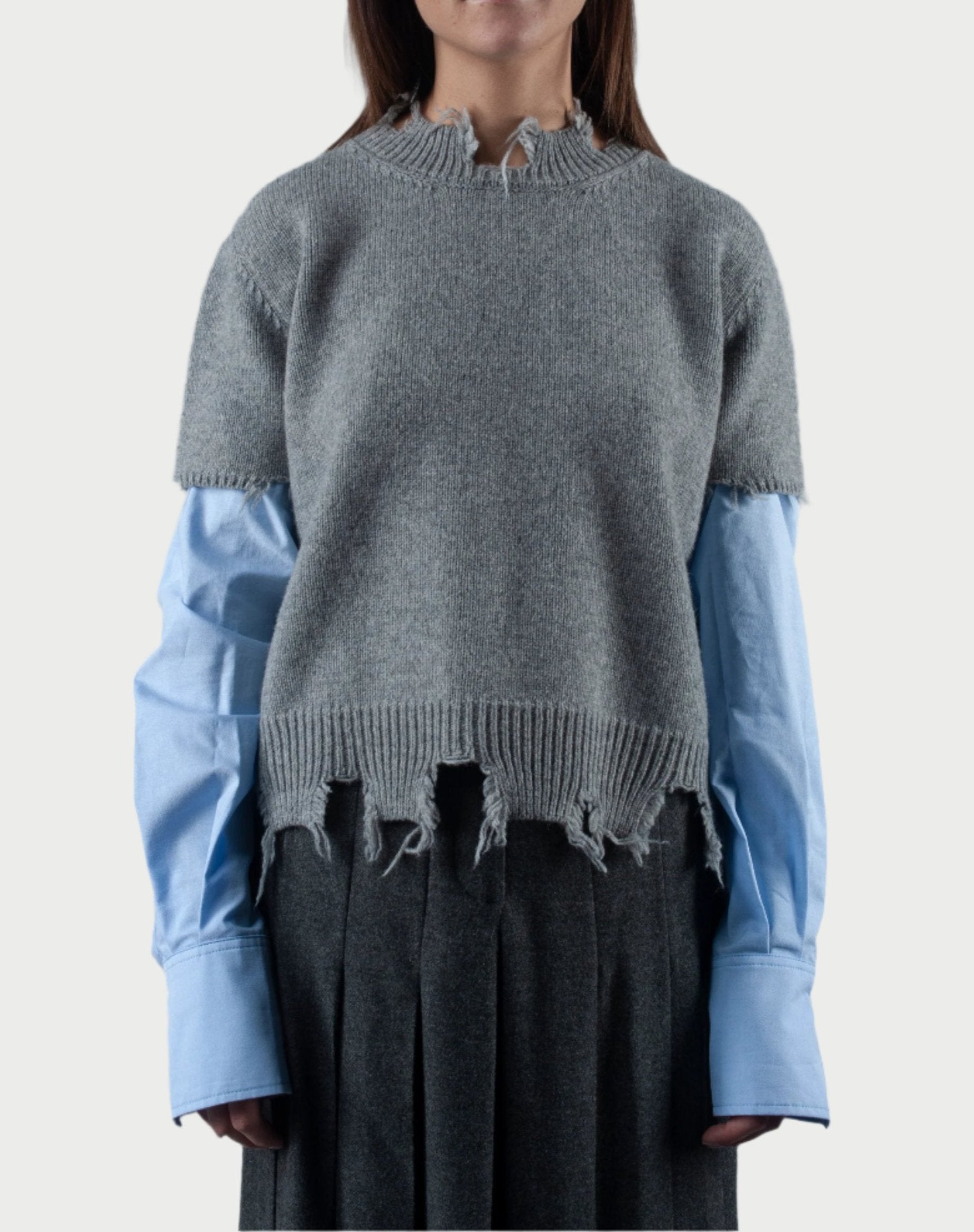 A person showcases a layered Phisique du role outfit with the Sweater Sleeves, featuring distressed gray detailing over a light blue shirt with cotton sleeves that extend beyond the frayed sweater cuffs. This ensemble is paired with pleated gray pants, embodying a stylish and effortlessly relaxed aesthetic.