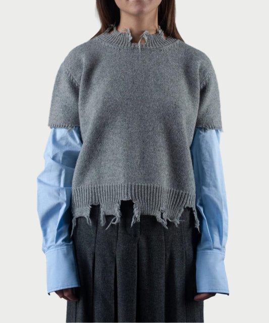 A person showcases a layered Phisique du role outfit with the Sweater Sleeves, featuring distressed gray detailing over a light blue shirt with cotton sleeves that extend beyond the frayed sweater cuffs. This ensemble is paired with pleated gray pants, embodying a stylish and effortlessly relaxed aesthetic.