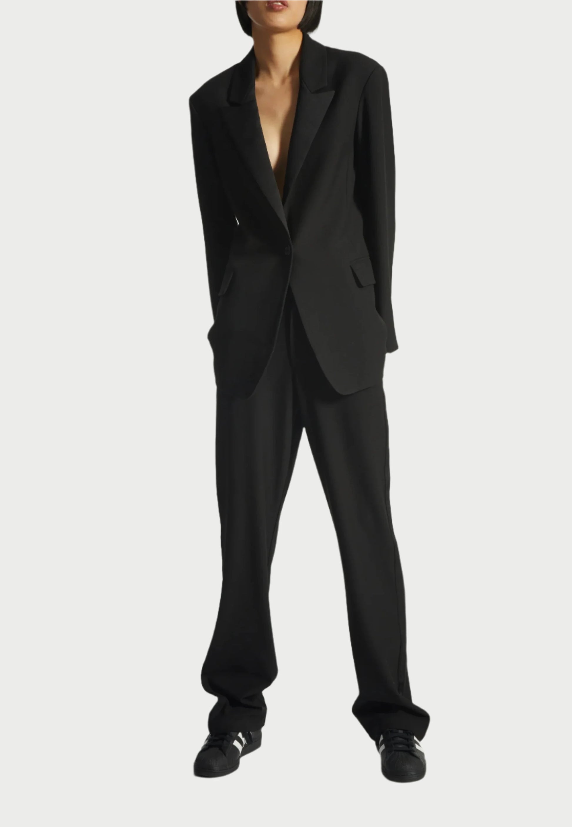 The individual sports a black oversized suit featuring a Saint Art NY Dylan Tuxedo Jacket, highlighted by charmeuse satin lapels. Paired with wide-leg pants and black sneakers, the relaxed pose has hands in pockets against a light grey backdrop.