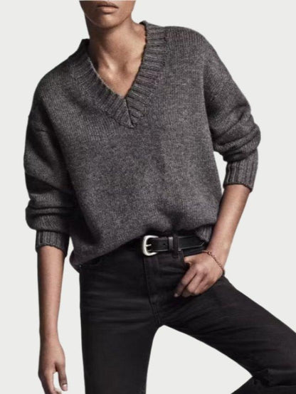 A person wearing a loose-fitting, grey Chunky V-neck Sweater by Frame tucked into black jeans with a black belt stands against a plain backdrop. They have white socks and black shoes, with one foot slightly raised. The overall look is casual and confident, perfect for the Fall season.