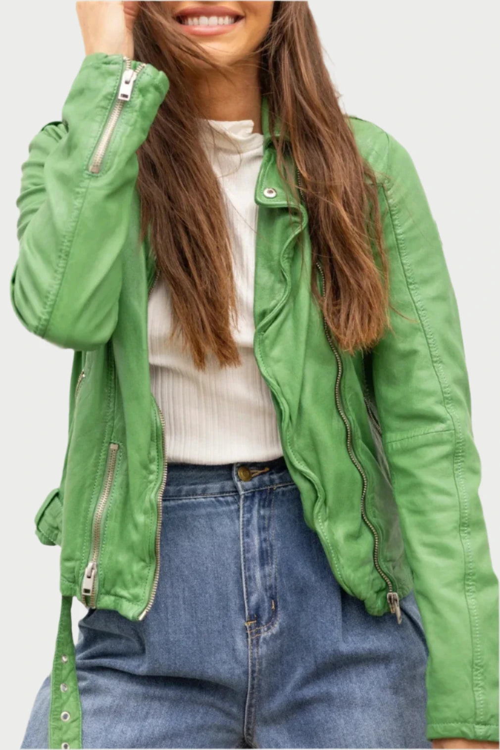 A person with long brown hair, smiling with their eyes closed, is wearing a snug fit green **Wild Moto Jacket** by **Mauritius** over a white top and blue denim jeans. Their left hand is brushing their hair back as they stand in front of a white textured wall.