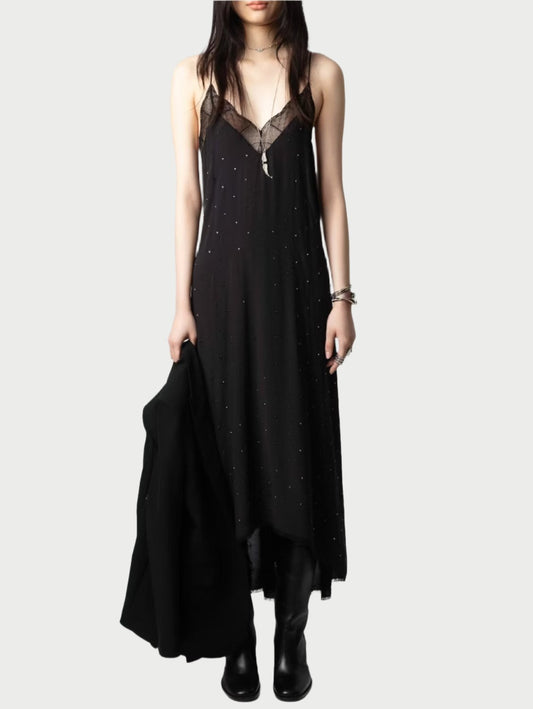 A person in a long black Zadig & Voltaire Risty Dress with sheer details and delicate straps holds a black coat. They accessorize with a long necklace and bracelets, completing the look with black boots against a plain light background.